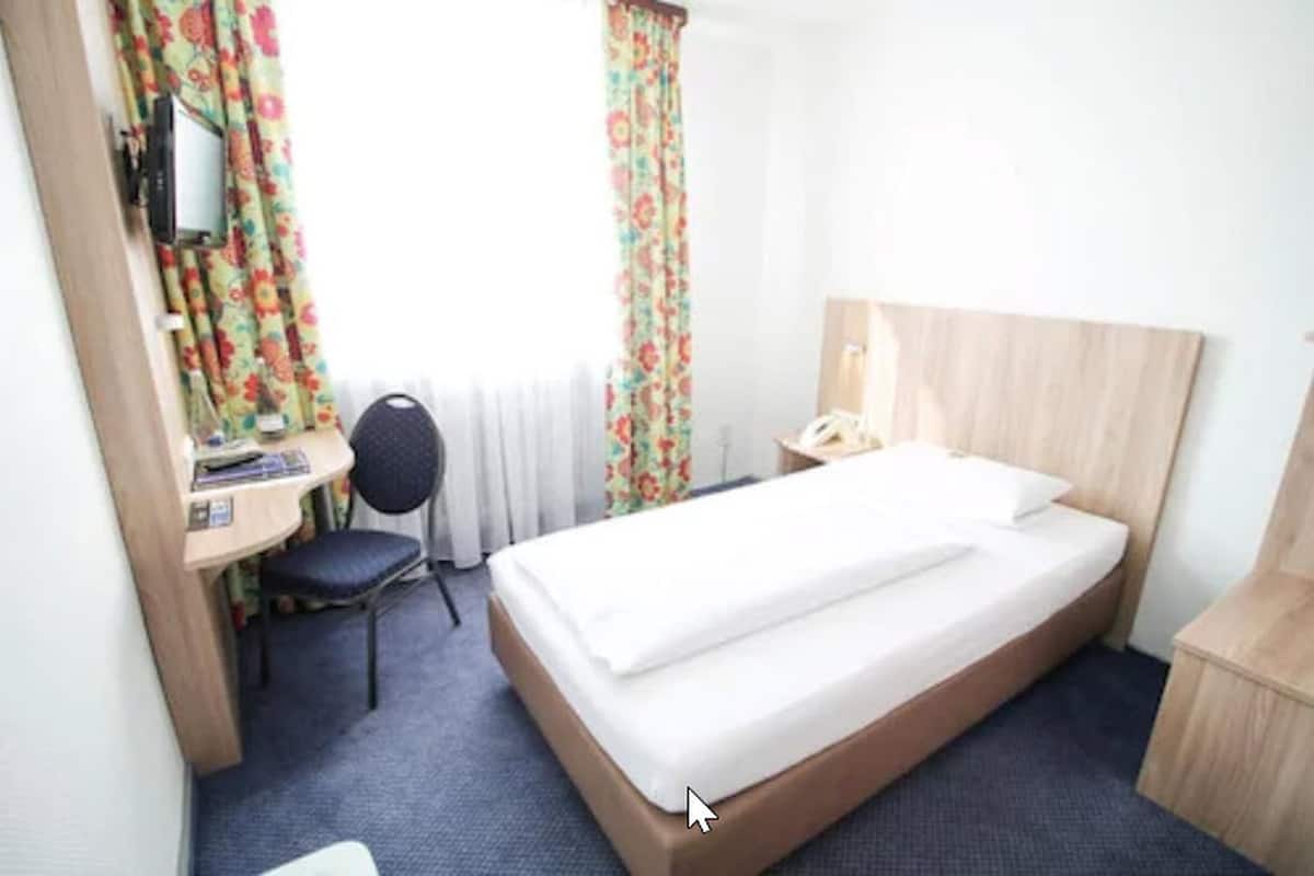 Una habitación de hotel con una cama individual, un escritorio con silla, una mesita de noche y un ventanal con cortinas florales.
