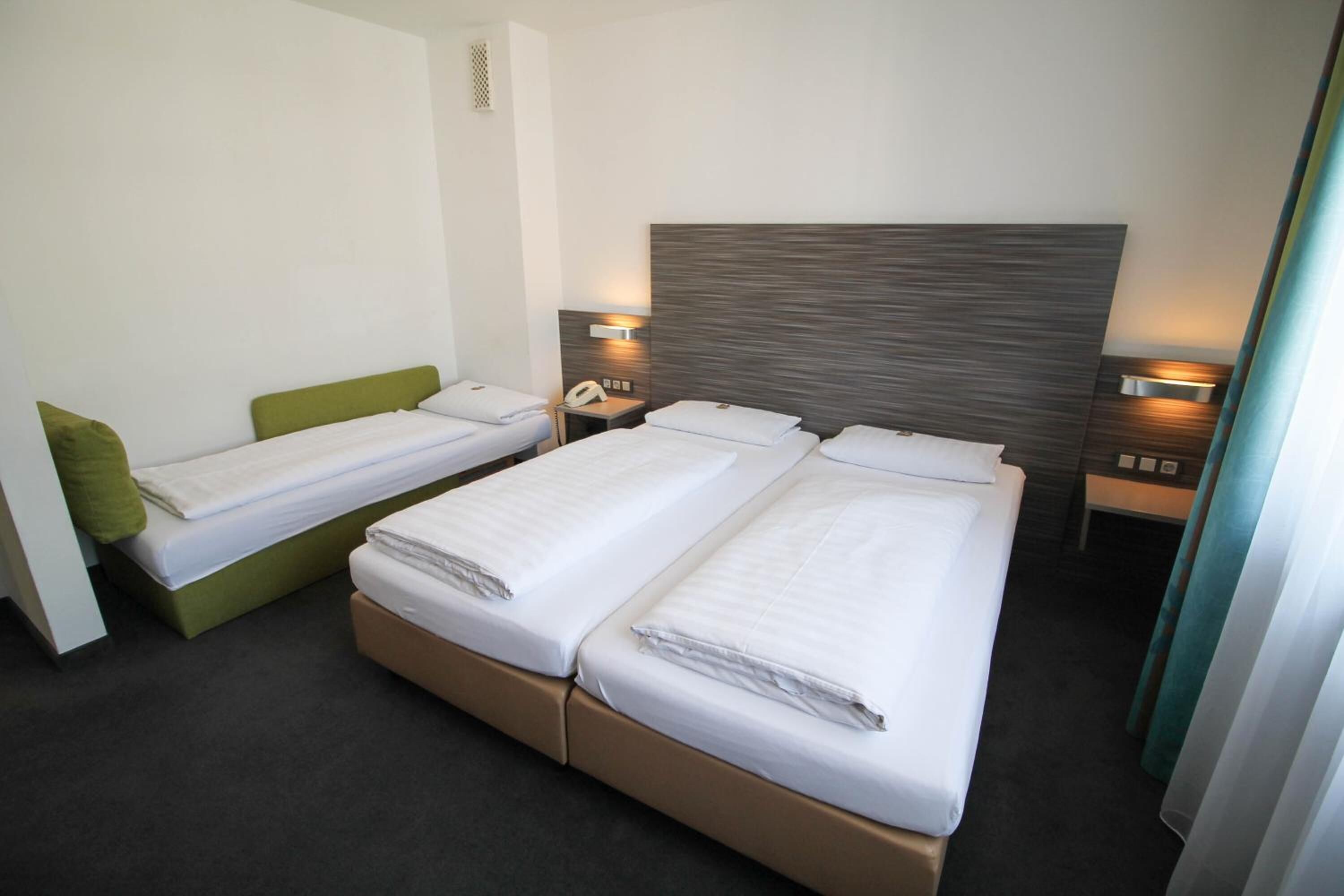 A hotel room with two single beds, a green accent bed, a large headboard, bedside lamps, and a telephone.