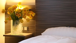 A hotel room with a bedside table holding a vase of yellow flowers, a nightstand with a lamp, and a bed with white bedding. - Hotel Am Zoo (Frankfurt)
