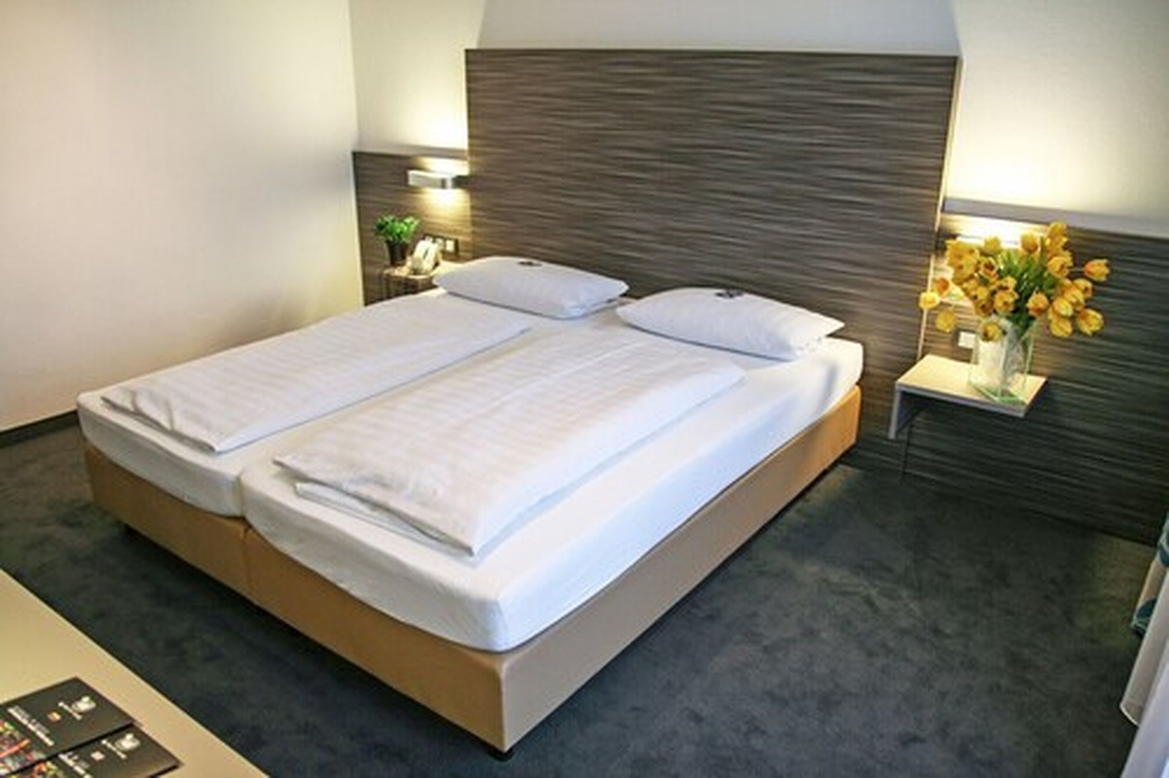 A double bed with a wooden headboard, a bedside table with a lamp, and a vase with flowers.