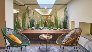 A modern interior with a seating area featuring two green and two brown wicker chairs, a small round table with a magazine, and a wooden bench.