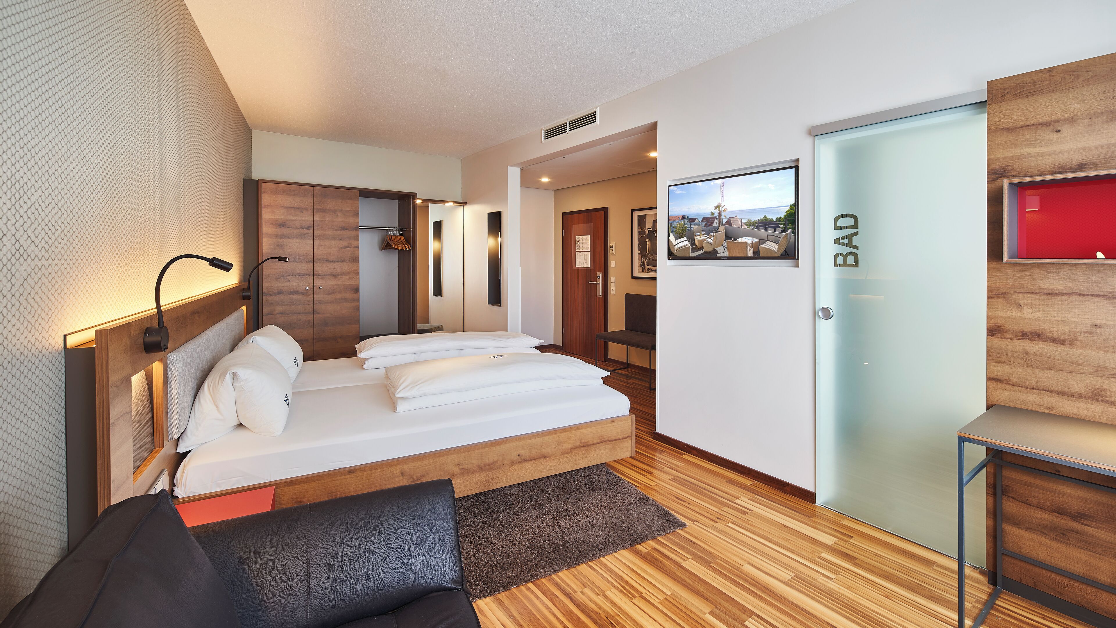 A hotel room with a wooden bed, a desk, a chair, and a TV on the wall.