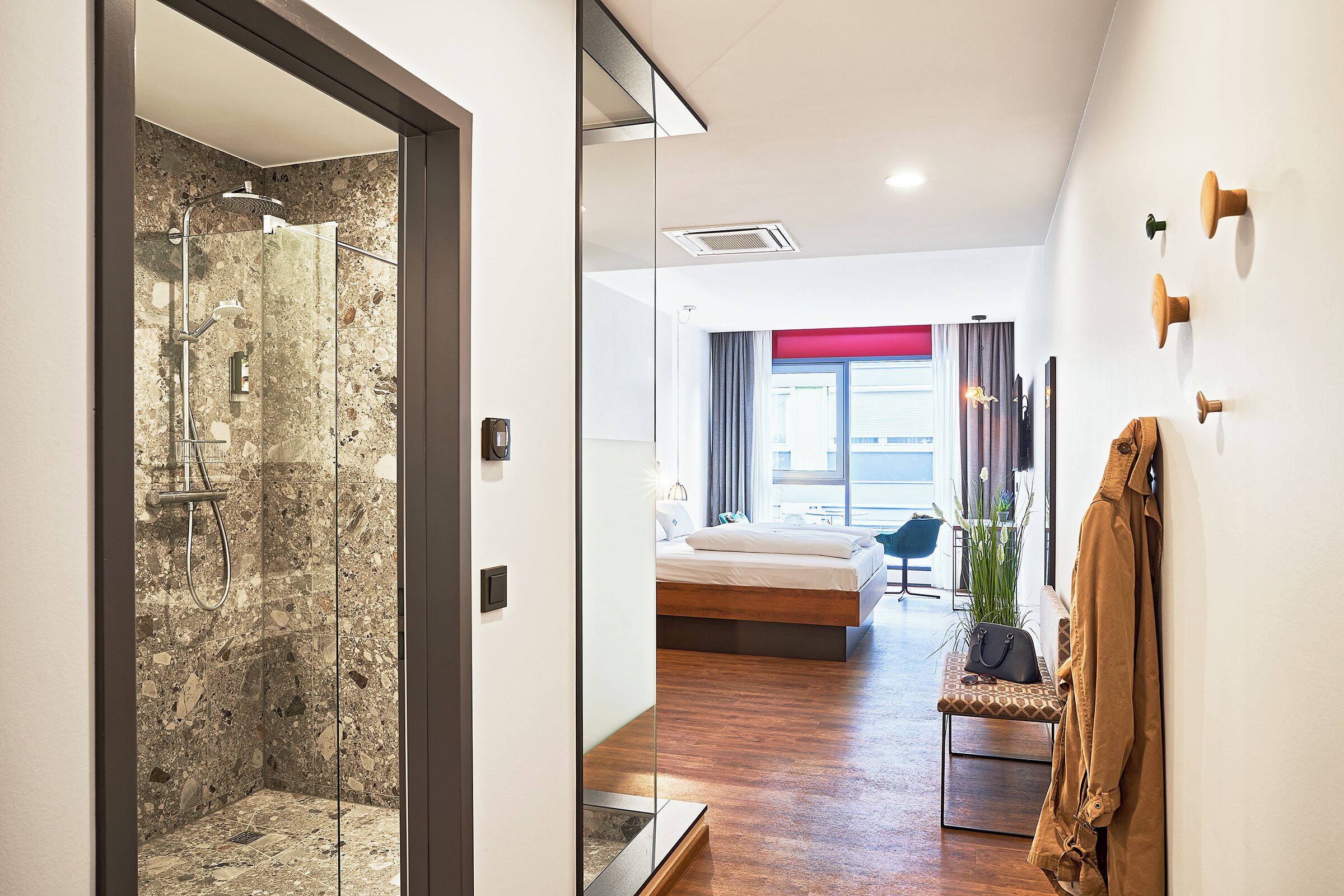 A modern hotel room with a glass shower enclosure, a wooden floor, a bed with a red accent wall, and a coat hanging on the wall.