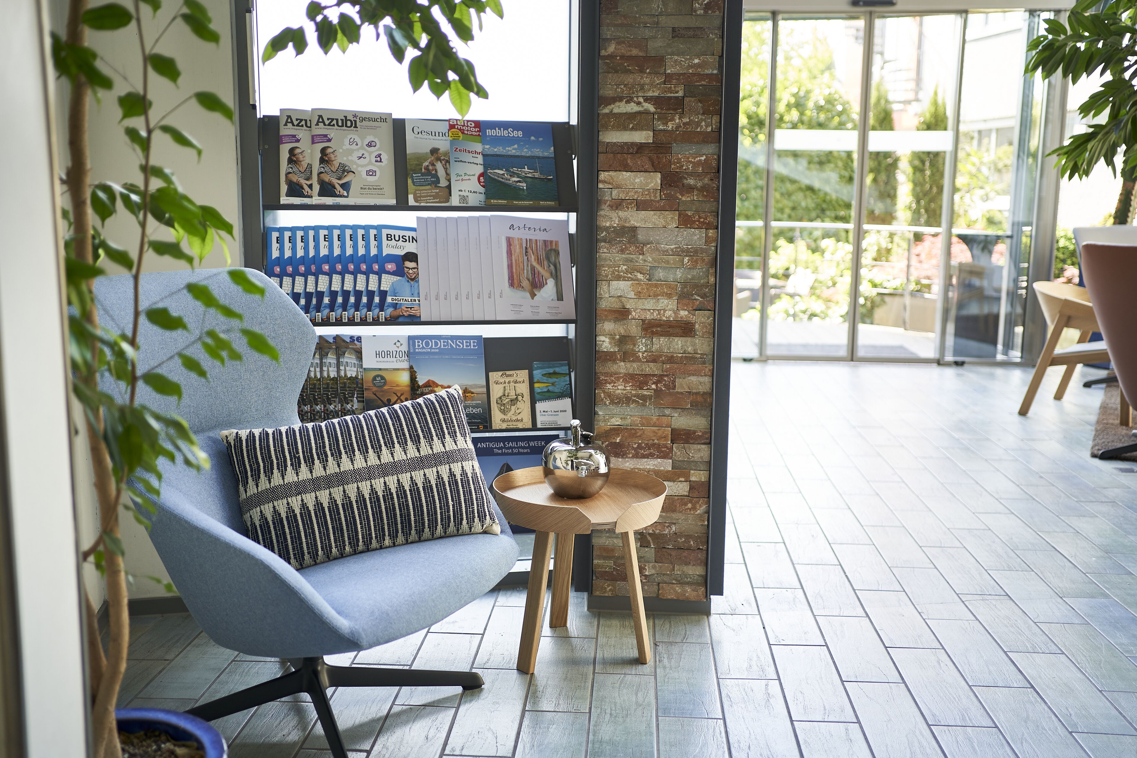 Ein moderner Lounge-Bereich mit einem blauen Sessel, einem kleinen Holztisch mit Teekanne sowie einem Bücherregal mit Zeitschriften und Büchern.