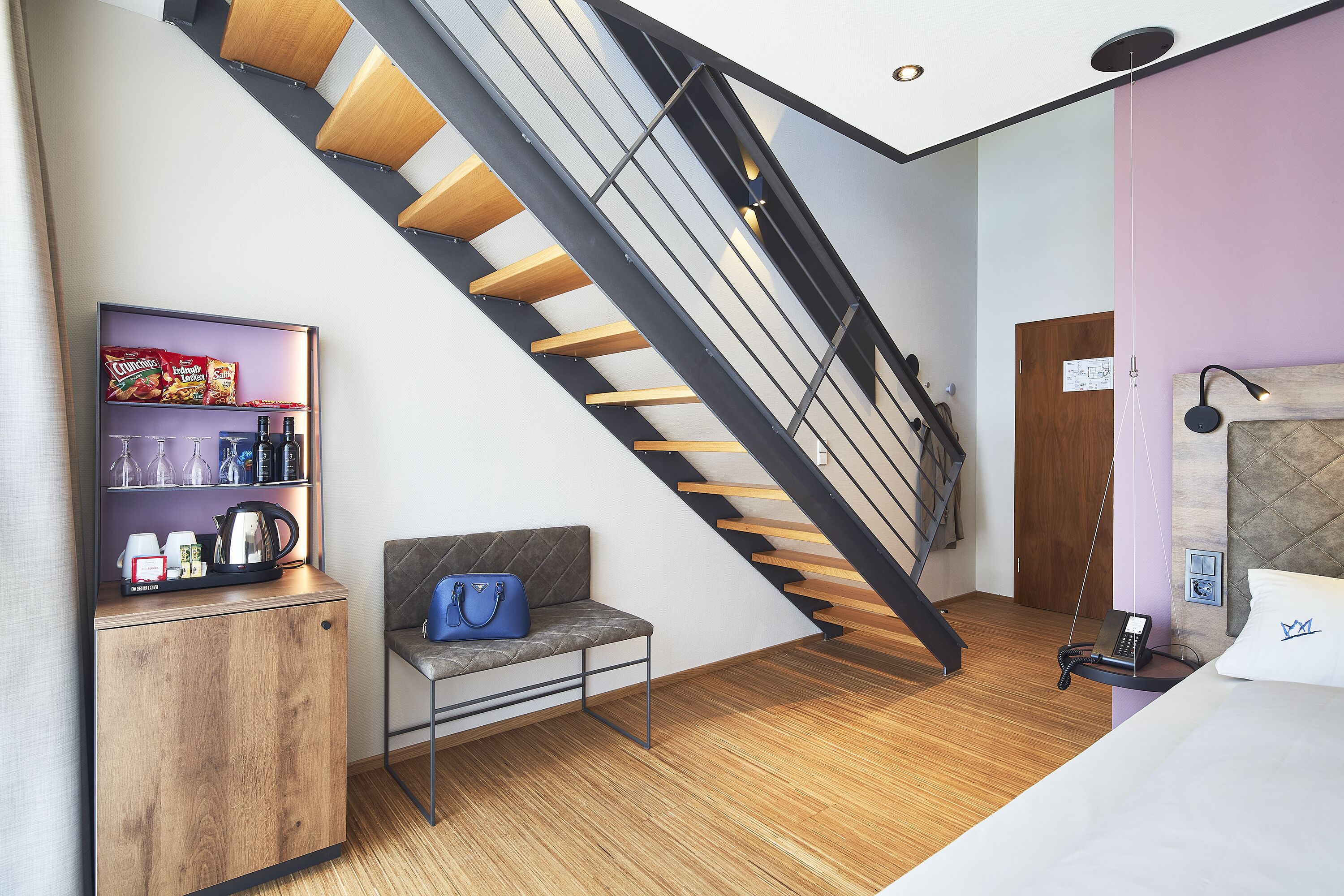 Een moderne hotelkamer met een houten vloer, een grijze bank, een klein houten kastje met snacks, een blauwe handtas, een trap met zwarte leuningen en een bed met een wit kussen.