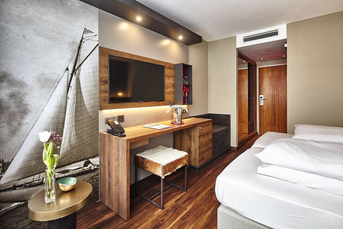 A modern hotel room with a wooden desk, a large mirror, a bed, and a small stool with a vase of flowers.