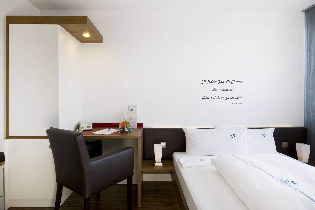 A hotel room with a bed, a desk, and a chair. There is a wall with a quote in German.