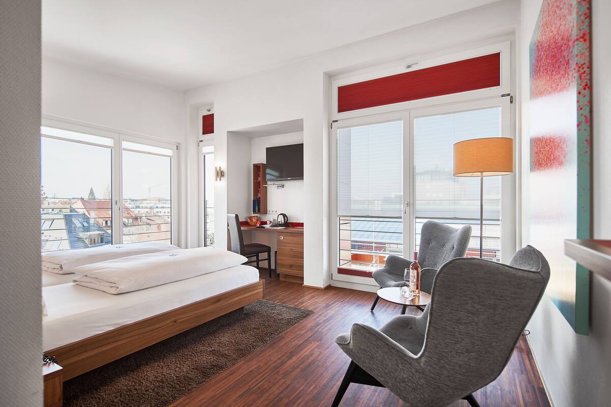 A modern hotel room with a bed, desk, and seating area, large windows, and a view of the city.