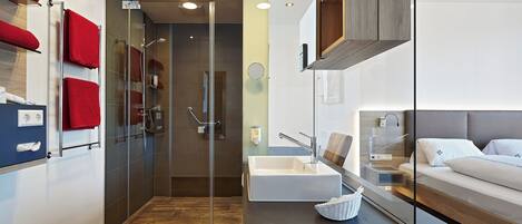 Un baño moderno con cabina de ducha de vidrio, un lavamanos blanco y una mesita de noche con una cesta.