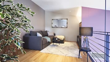 A modern living room with a grey sofa, a small side table, a potted plant, and a large mirror on the wall.