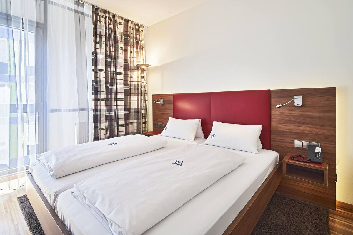 A double bed with white linens and a red headboard, a bedside table with a phone, and a window with sheer curtains.