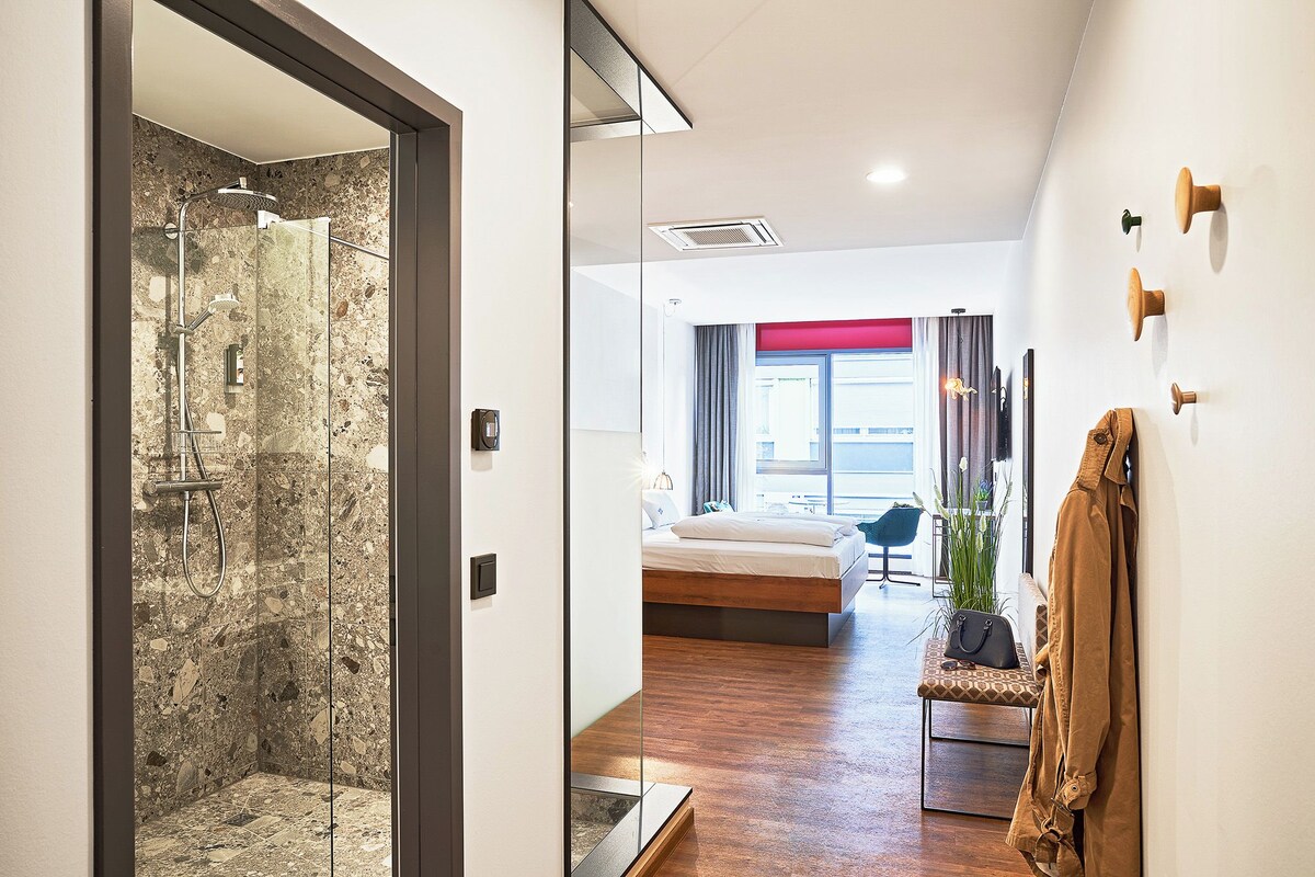 Habitación de hotel moderna con cabina de ducha de vidrio, cama con pared de acento roja, banco con un bolso y abrigo colgado en la pared.