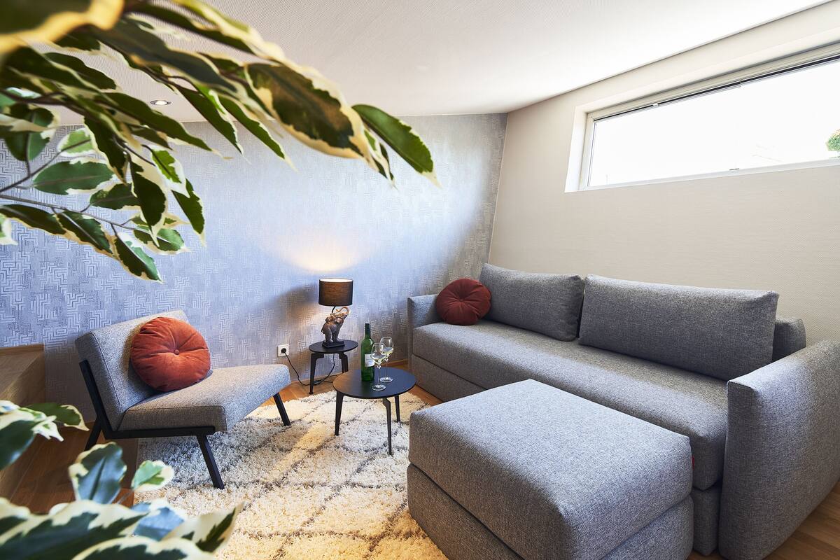 A modern living room with a grey sofa, a matching armchair, and a small side table with a lamp and wine glasses.