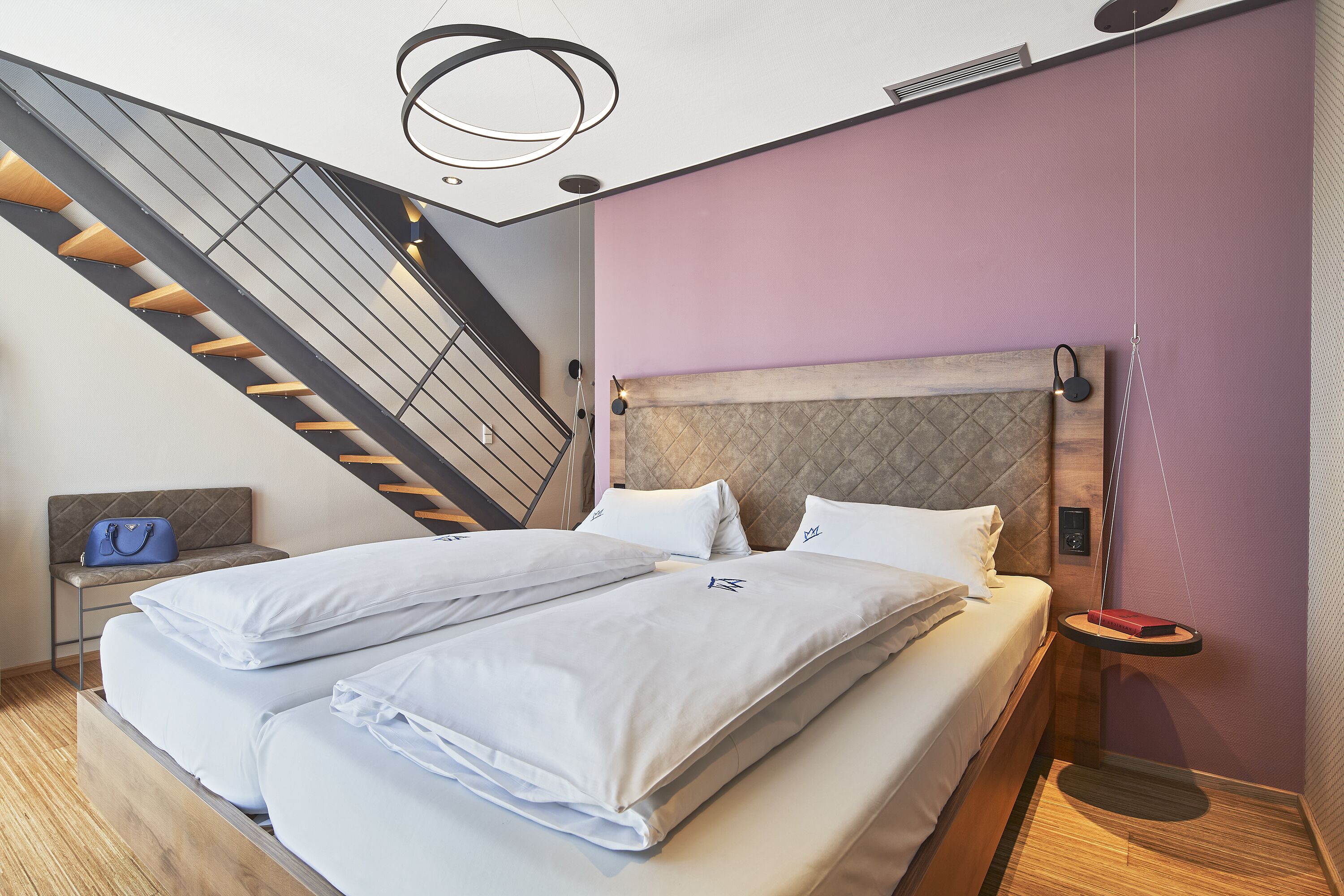 Una habitación de hotel moderna con una cama grande, un cabecero con diseño acolchado, una mesita de noche pequeña con una lámpara, y una escalera al fondo.