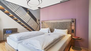 Una habitación de hotel moderna con una cama grande, un cabecero con diseño acolchado, una mesita de noche pequeña con una lámpara, y una escalera al fondo.