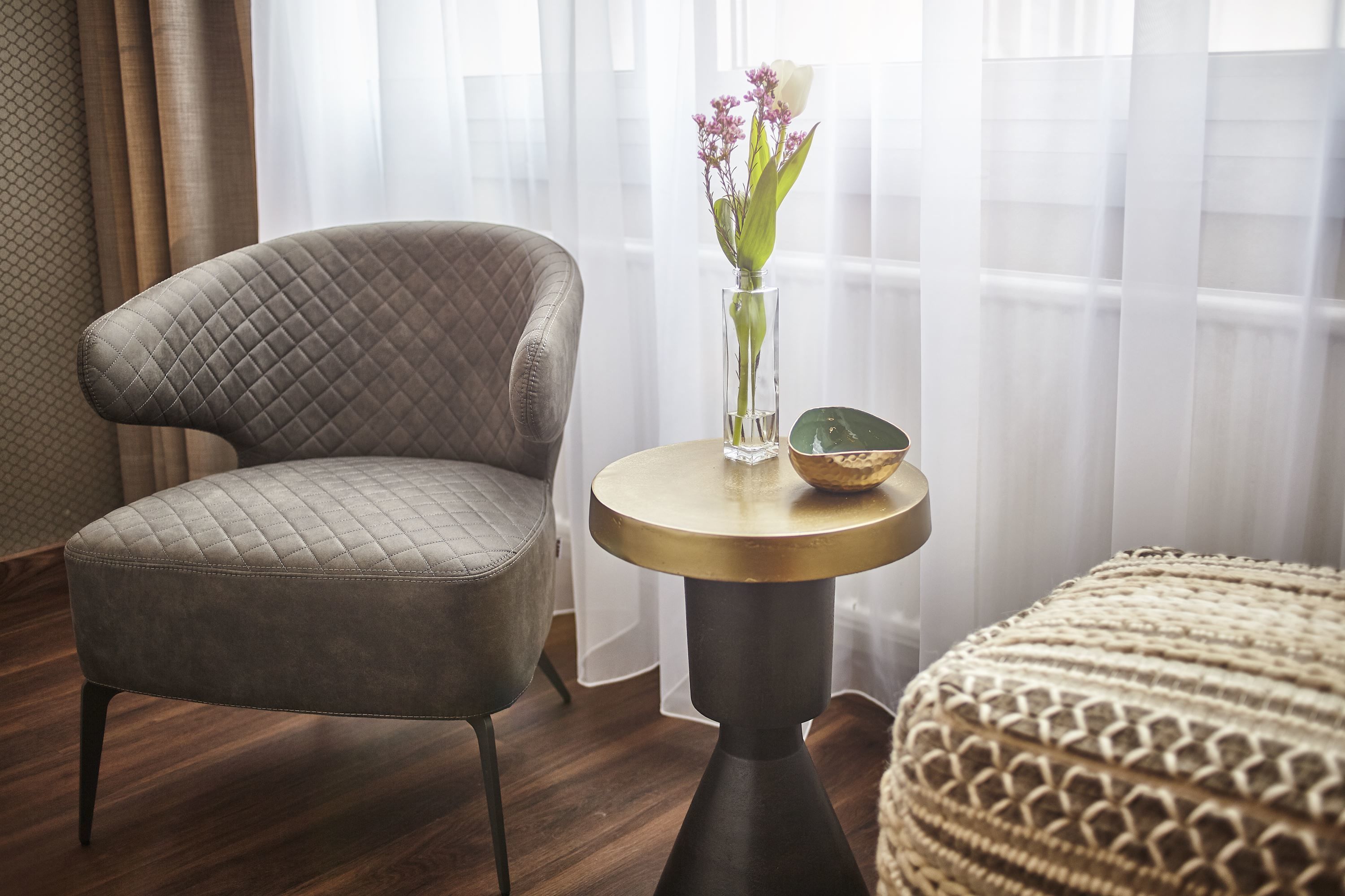 A room with a tufted armchair, a round table with a vase of flowers, and a textured ottoman.
