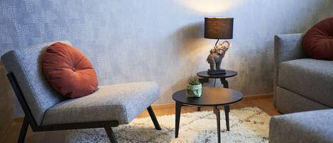 Un salón con un sofá gris, un sillón a juego, una mesita redonda con una lámpara y un papel tapiz estampado.