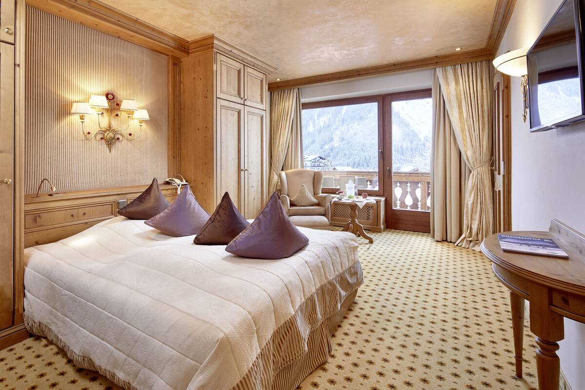Une chambre d’hôtel avec un grand lit, une tête de lit en bois, un lustre, un balcon offrant une vue sur les montagnes et une petite table avec une lampe.