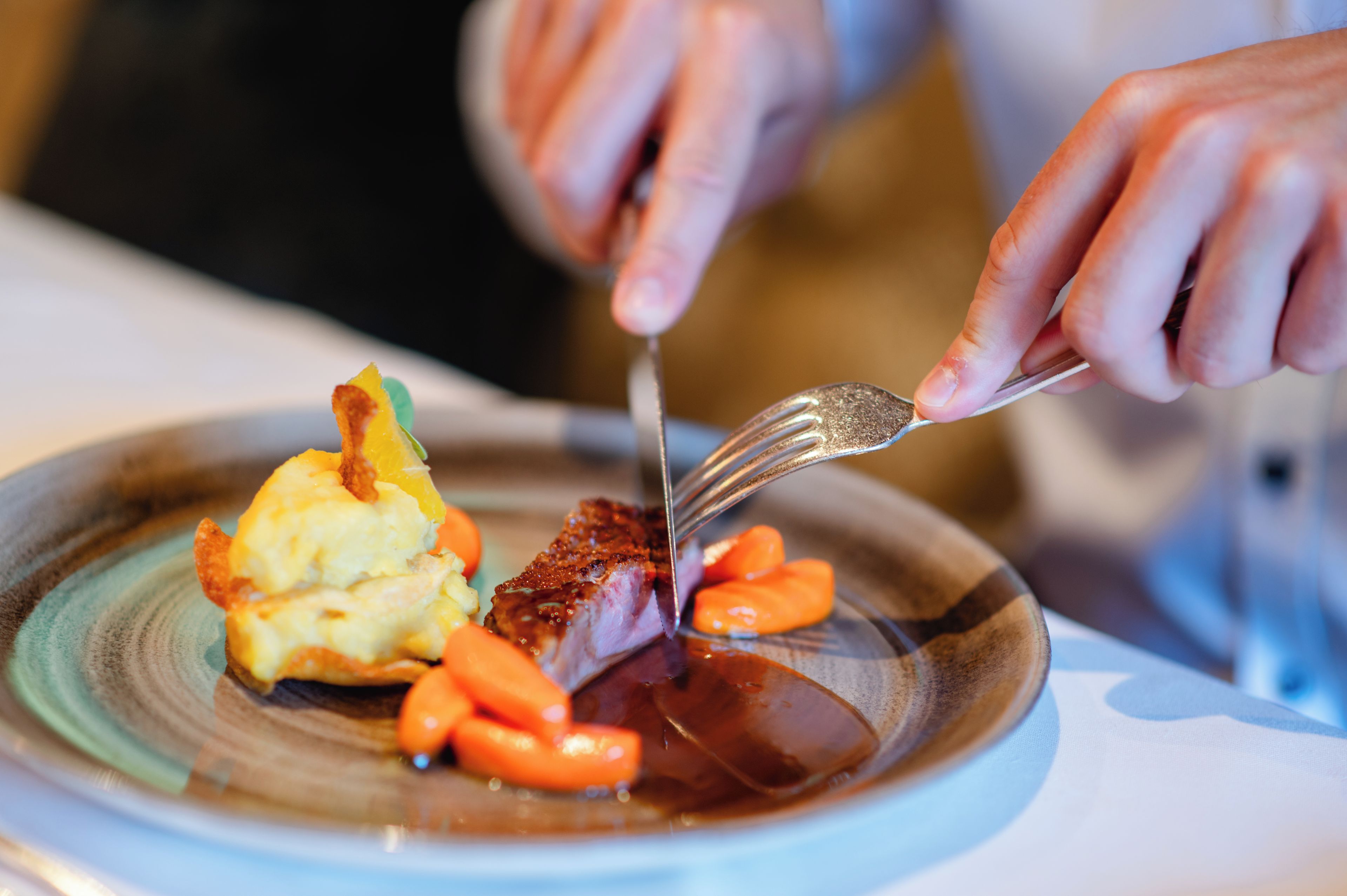 A person cutting a piece of meat with a fork and knife on a plate with vegetables.