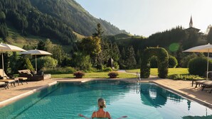 Una piscina cristalina con una persona nadando, rodeada de sillas de descanso y sombrillas, y un paisaje montañoso al fondo.
