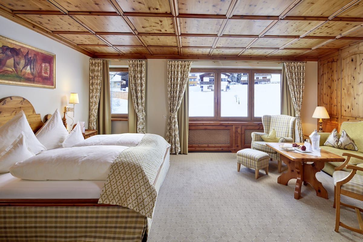 Une chambre d’hôtel avec un plafond en bois, un lit recouvert de draps blancs, un canapé vert, une table en bois et une vue sur un paysage enneigé par de grandes fenêtres.