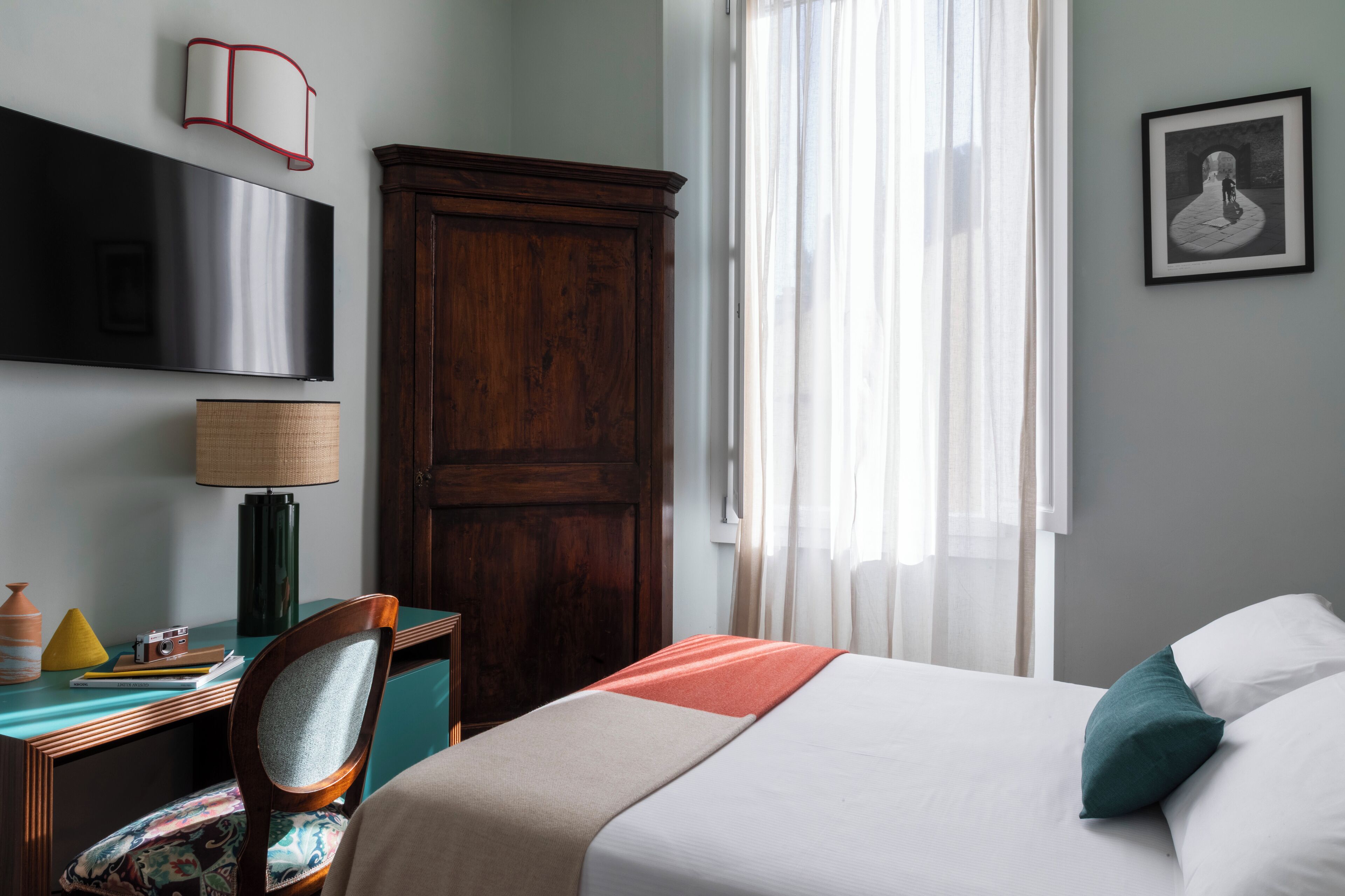 Une chambre d’hôtel comprenant un lit, un bureau avec une lampe, une chaise, une armoire, un téléviseur et une fenêtre avec des rideaux.