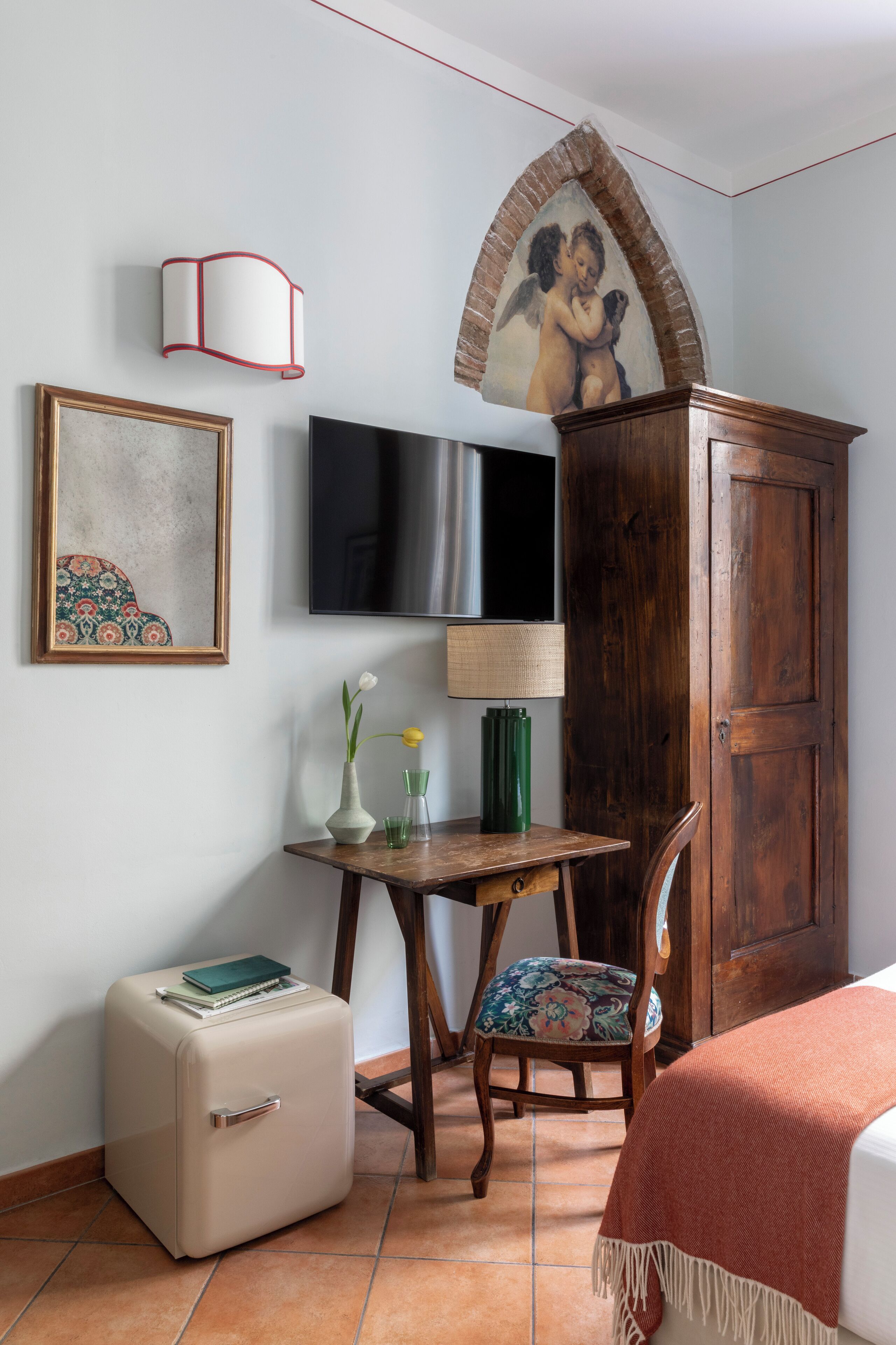 Um quarto com cama, um armário de madeira, televisão, quadro emoldurado, mesa pequena com abajur e um espelho decorativo.