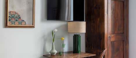 Um quarto com cama, um armário de madeira, televisão, quadro emoldurado, mesa pequena com abajur e um espelho decorativo.