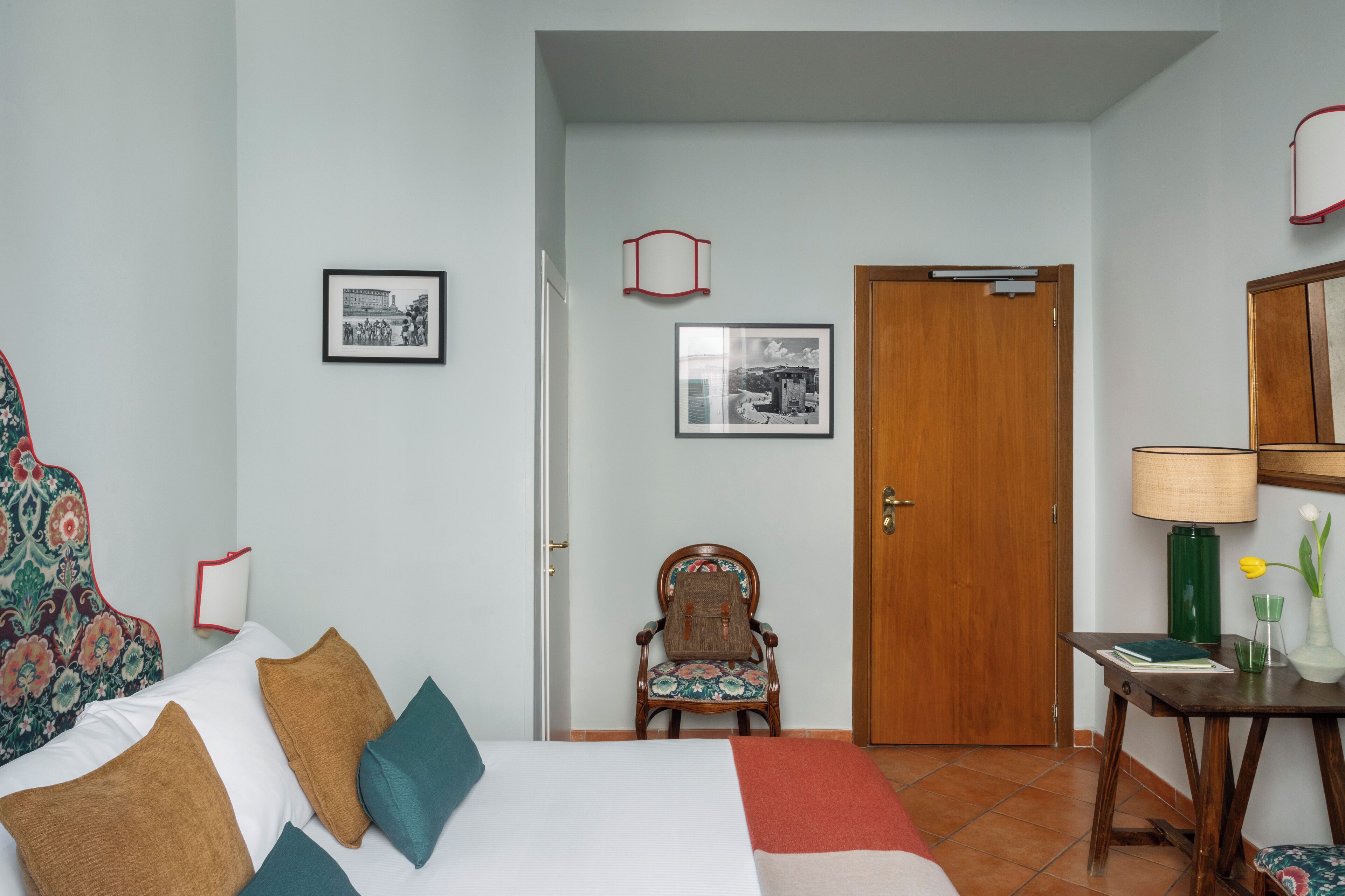 A hotel room with a bed, a chair, a desk with a lamp, and two framed pictures on the wall.