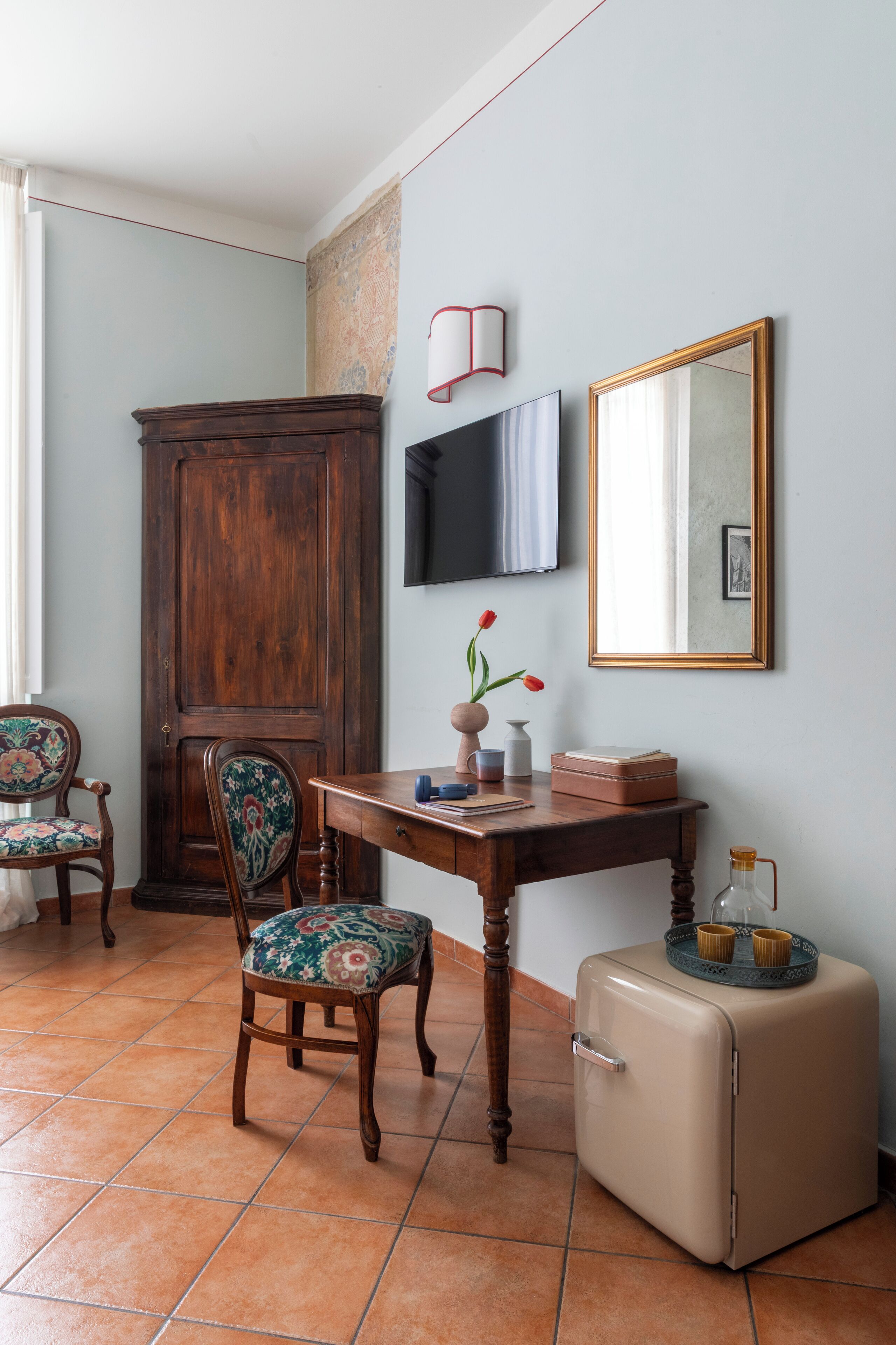 A room with a wooden cabinet, two chairs, a table with a vase and books, a television, a mirror, and a vintage suitcase.