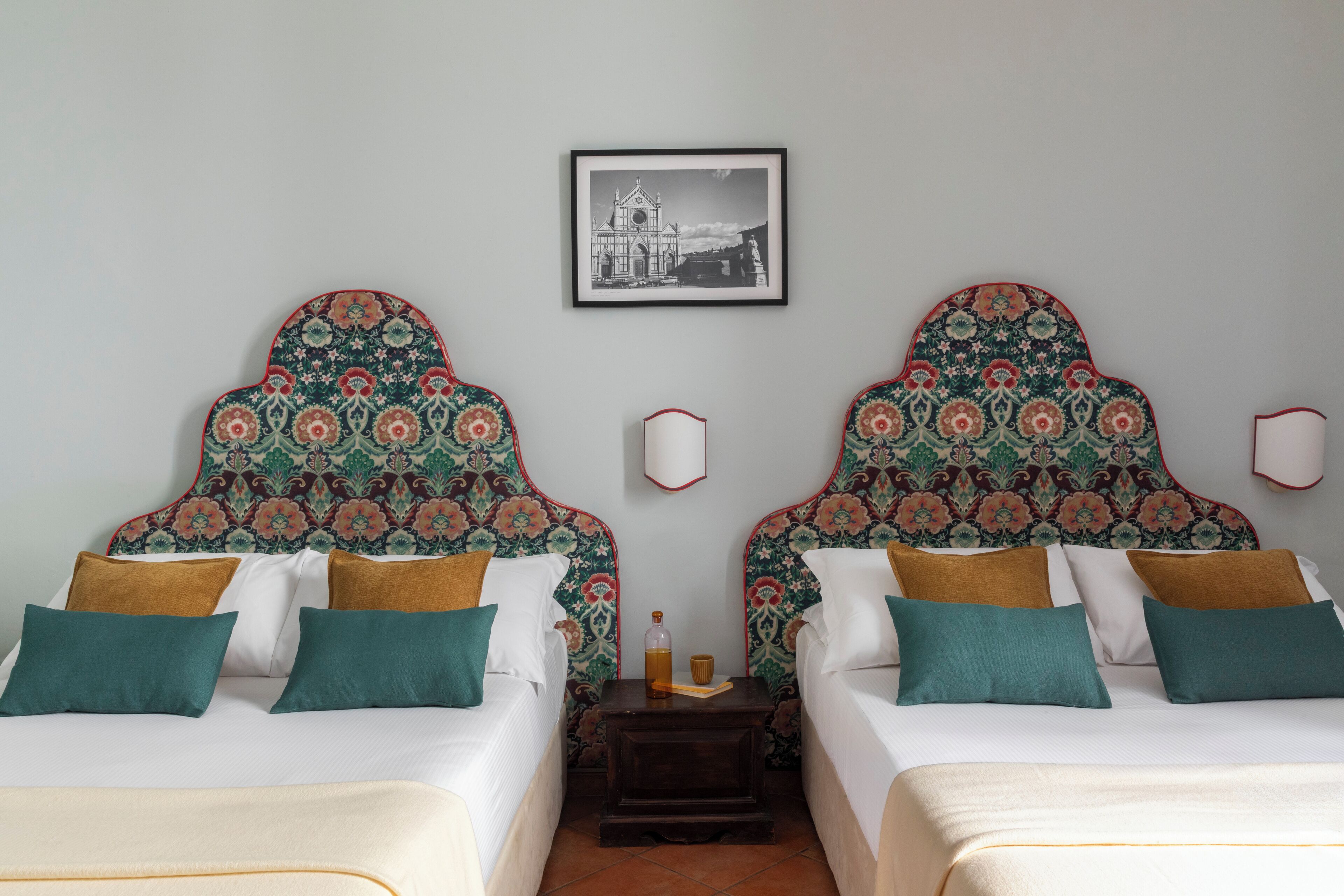 A hotel room with two beds, patterned headboards, white bedding, teal and brown pillows, a small table with a bottle and a cup, and a framed picture on the wall.