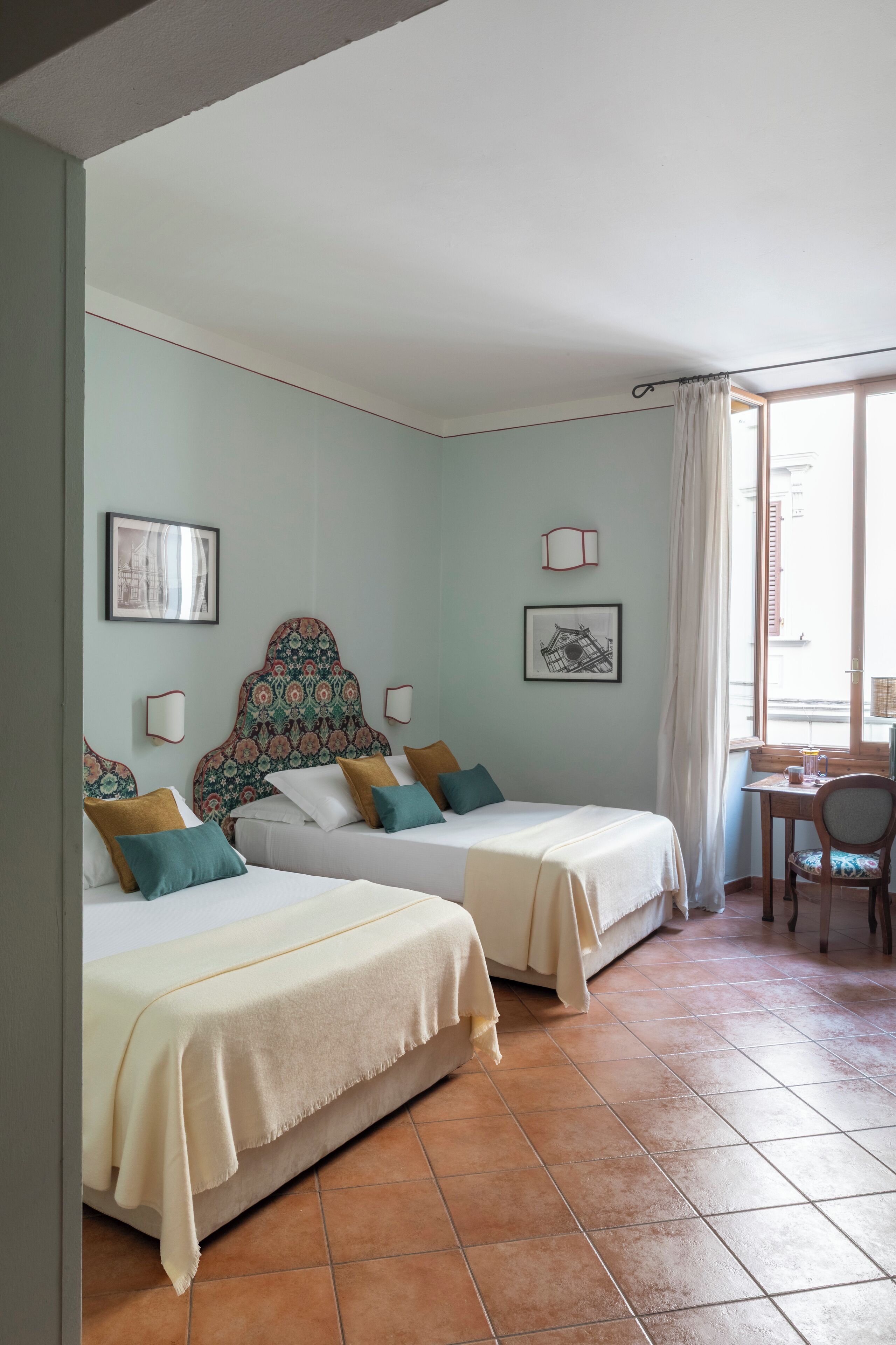 Ein Zimmer mit zwei Betten, einem Fenster mit Vorhängen, einem kleinen Tisch und einem Stuhl.