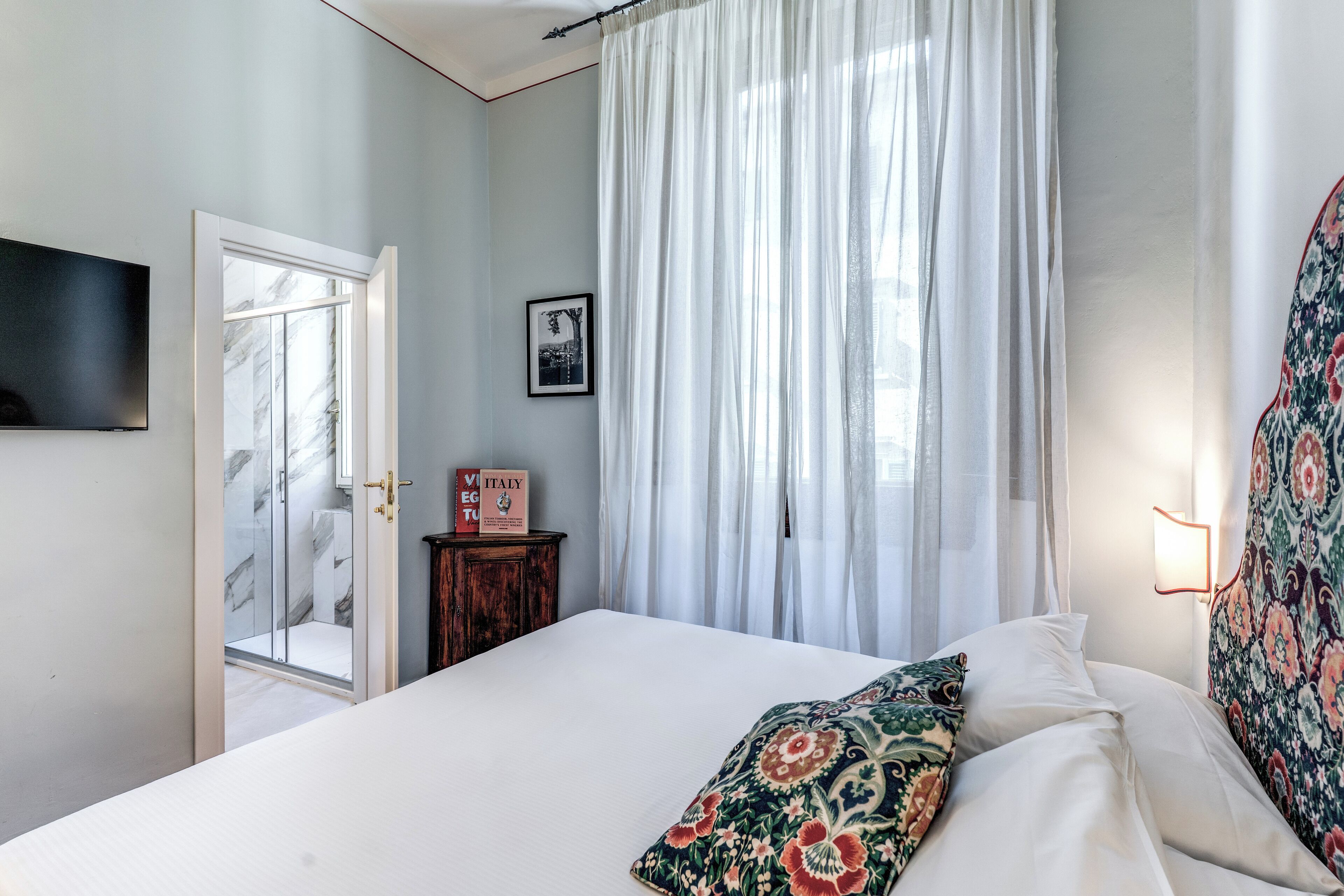 A bedroom with a bed, a shower, a door, a framed picture, and a wall-mounted light fixture.
