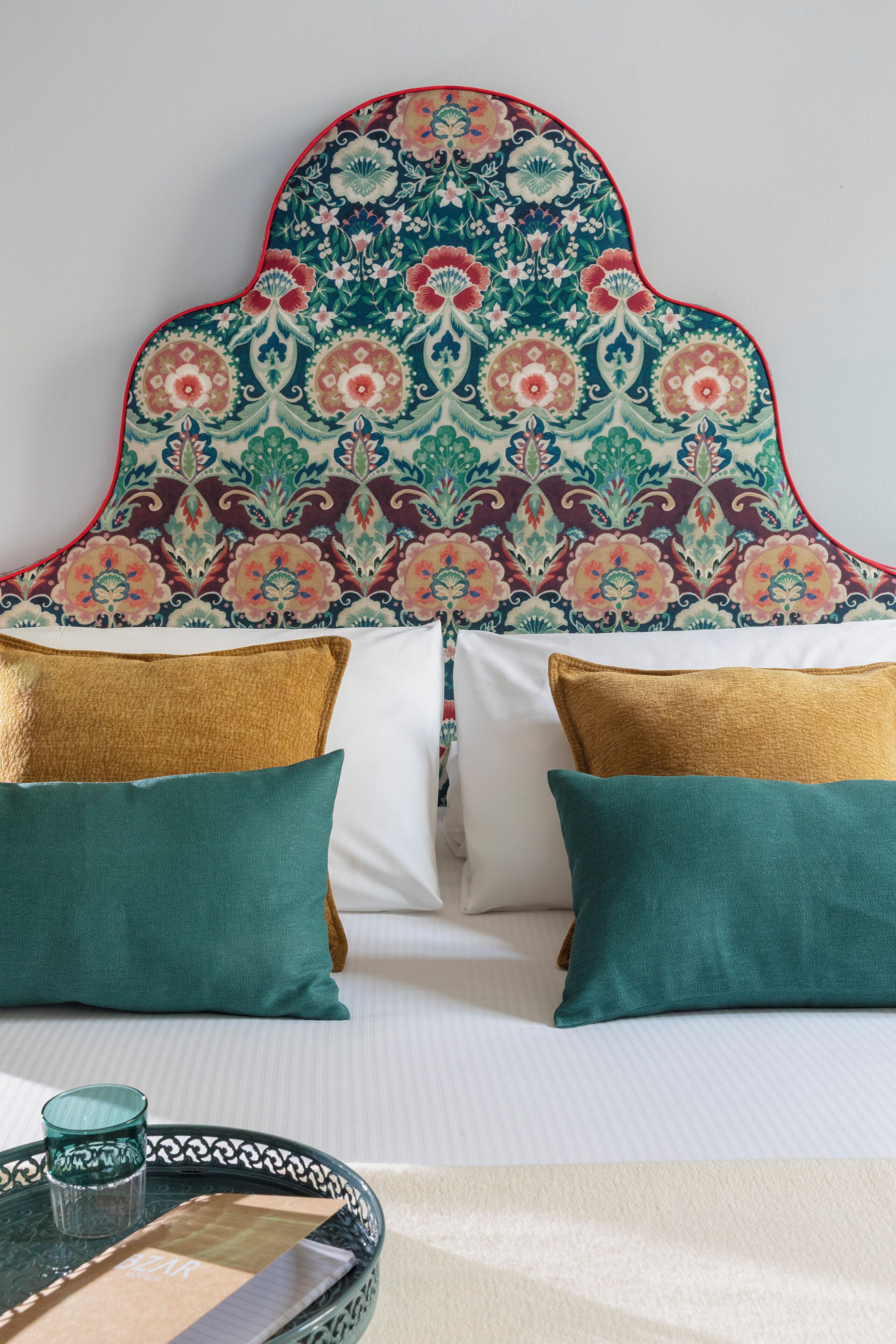 A bed with a patterned headboard, two gold and two green pillows, and a glass on a tray.