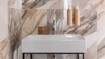 Un baño con pared de mármol, un espejo redondo, un lavamanos blanco, un marco metálico negro y dos botellas de crema corporal.