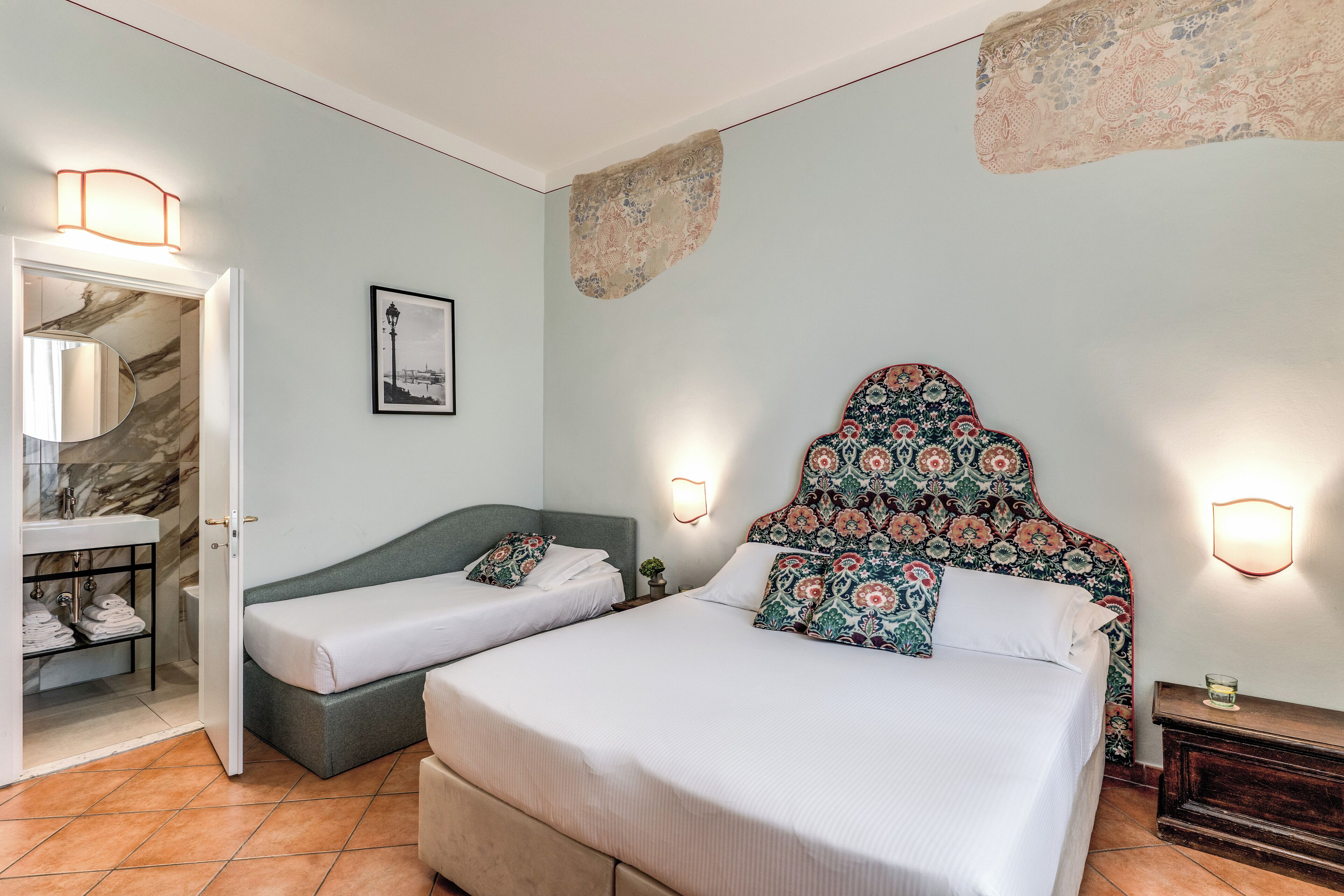 Une chambre d’hôtel comprenant un lit, une table de chevet, une commode, une lampe fixée au mur, un tableau encadré et une salle de bain avec un lavabo et un porte-serviettes.