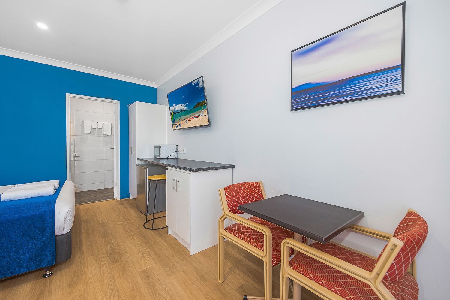 A compact room with a blue wall, a bed, a small kitchenette, and a dining area with a table and chairs.