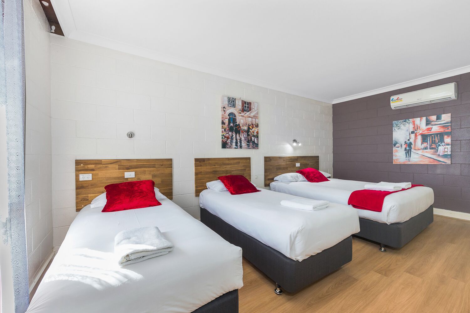 Habitació d'hotel amb tres llits individuals, roba de llit blanca, almoines vermelles i un capçal de llit gris.