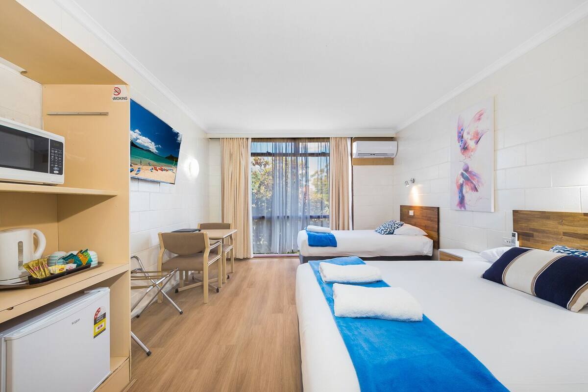 Quarto de hotel com duas camas, uma escrivaninha com micro-ondas, geladeira e uma janela grande com cortinas.