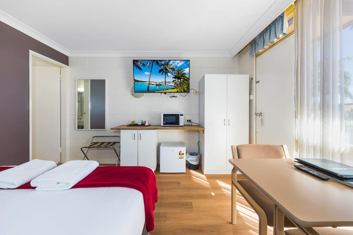 Quarto de hotel compacto com cama, escrivaninha, pequeno espaço de cozinha e televisão exibindo uma paisagem tropical.