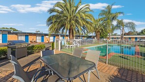 A poolside area with a glass table and chairs, a barbecue grill, and a fenced swimming pool. - Plainsman Motel (Forbes)