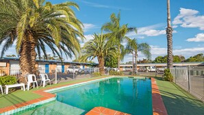 A swimming pool with palm trees, a fenced area, and a building with blue doors in the background. - Plainsman Motel (Forbes)
