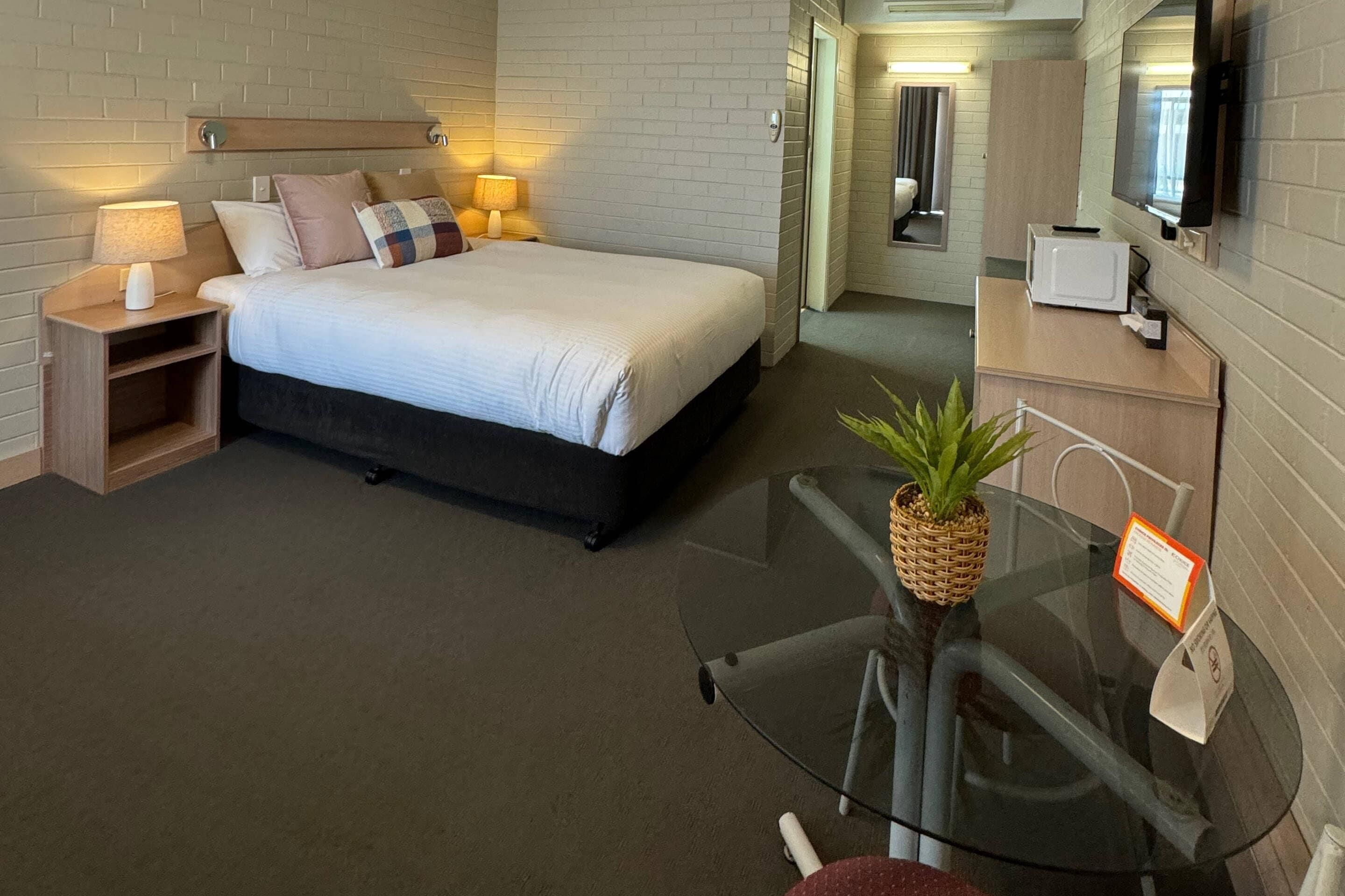 A hotel room with a bed, bedside lamps, a TV, a microwave, and a glass table with a plant.