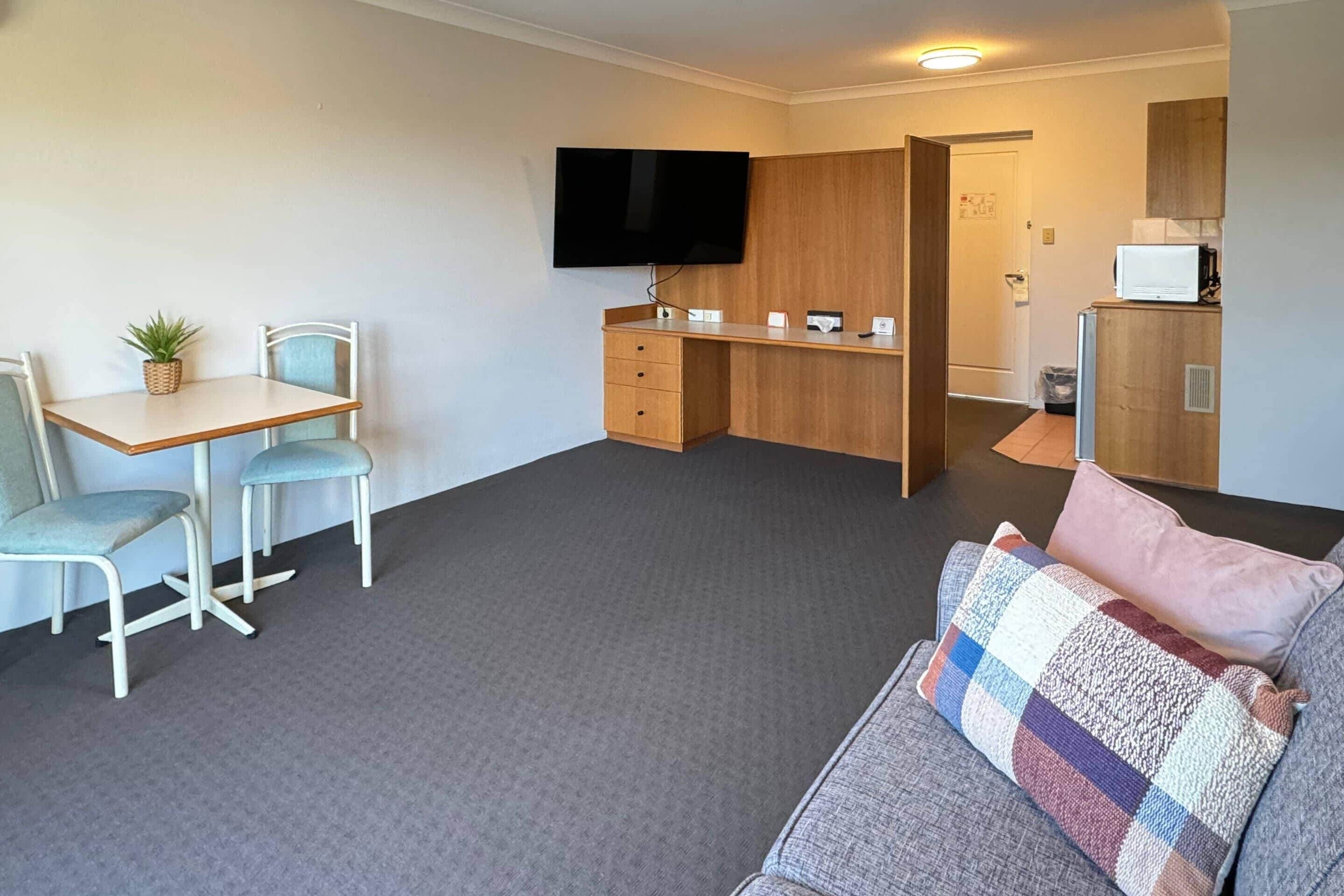 A compact room with a sofa, a small desk with a chair, a television, and a kitchenette area.