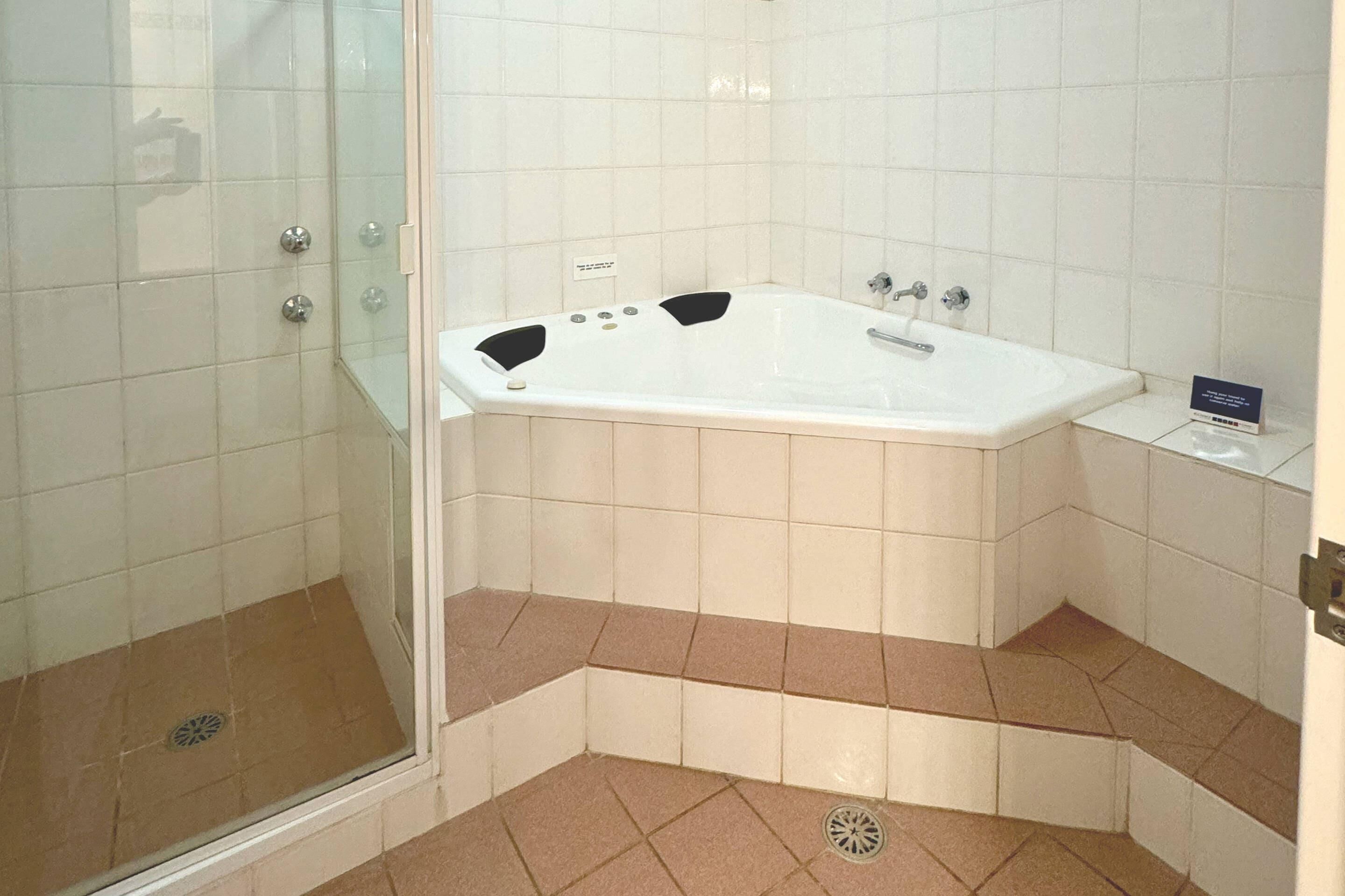 A bathroom with a glass shower enclosure, a white bathtub, and beige-tiled flooring.