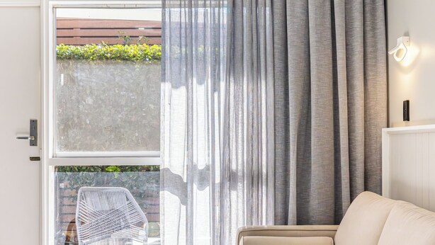 Ein modernes Hotelzimmer mit einem großen Bett, einer Couch, einem kleinen runden Tisch und Blick auf einen Garten durch eine Schiebetür.