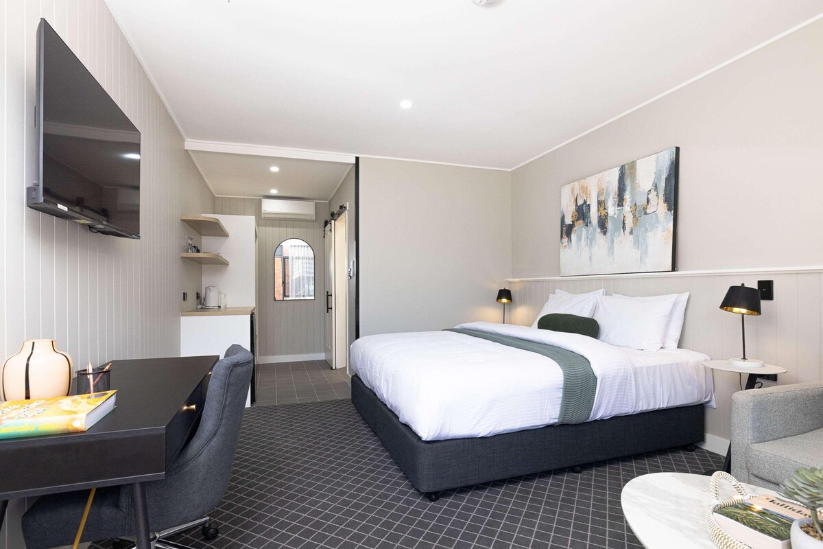 A hotel room with a bed, a desk, a chair, a TV, a painting, and a small table with a plant.