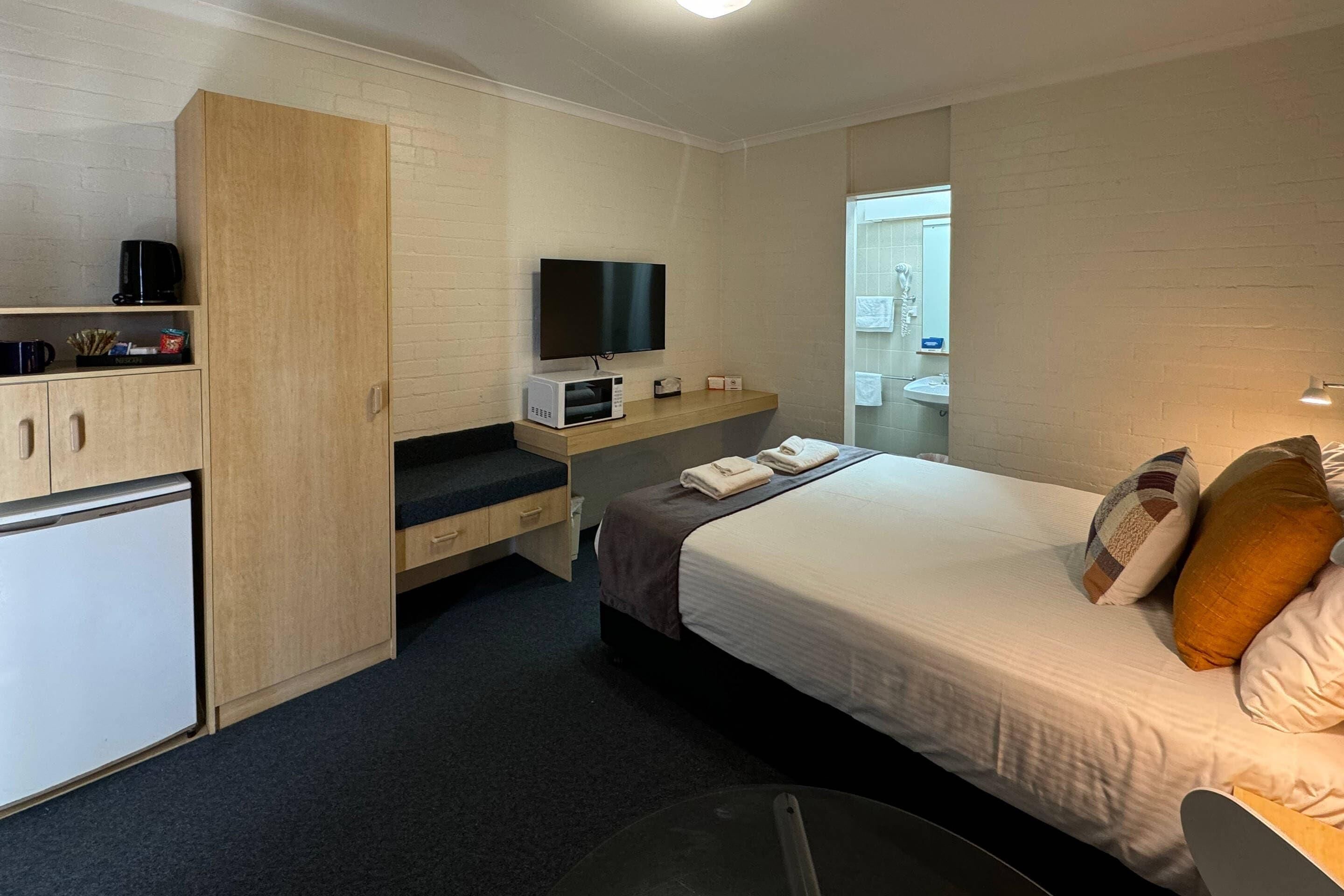 A hotel room with a bed, a TV, a microwave, a small fridge, a desk, and a bathroom visible through an open door.