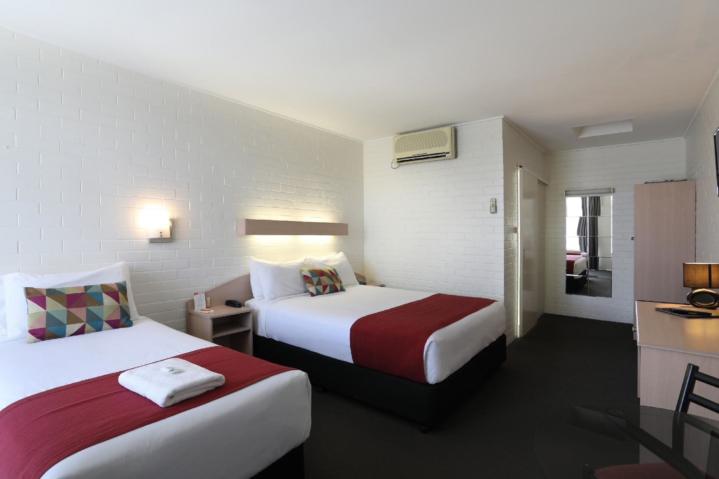 A hotel room with two beds, white brick walls, a desk, and a mirror.