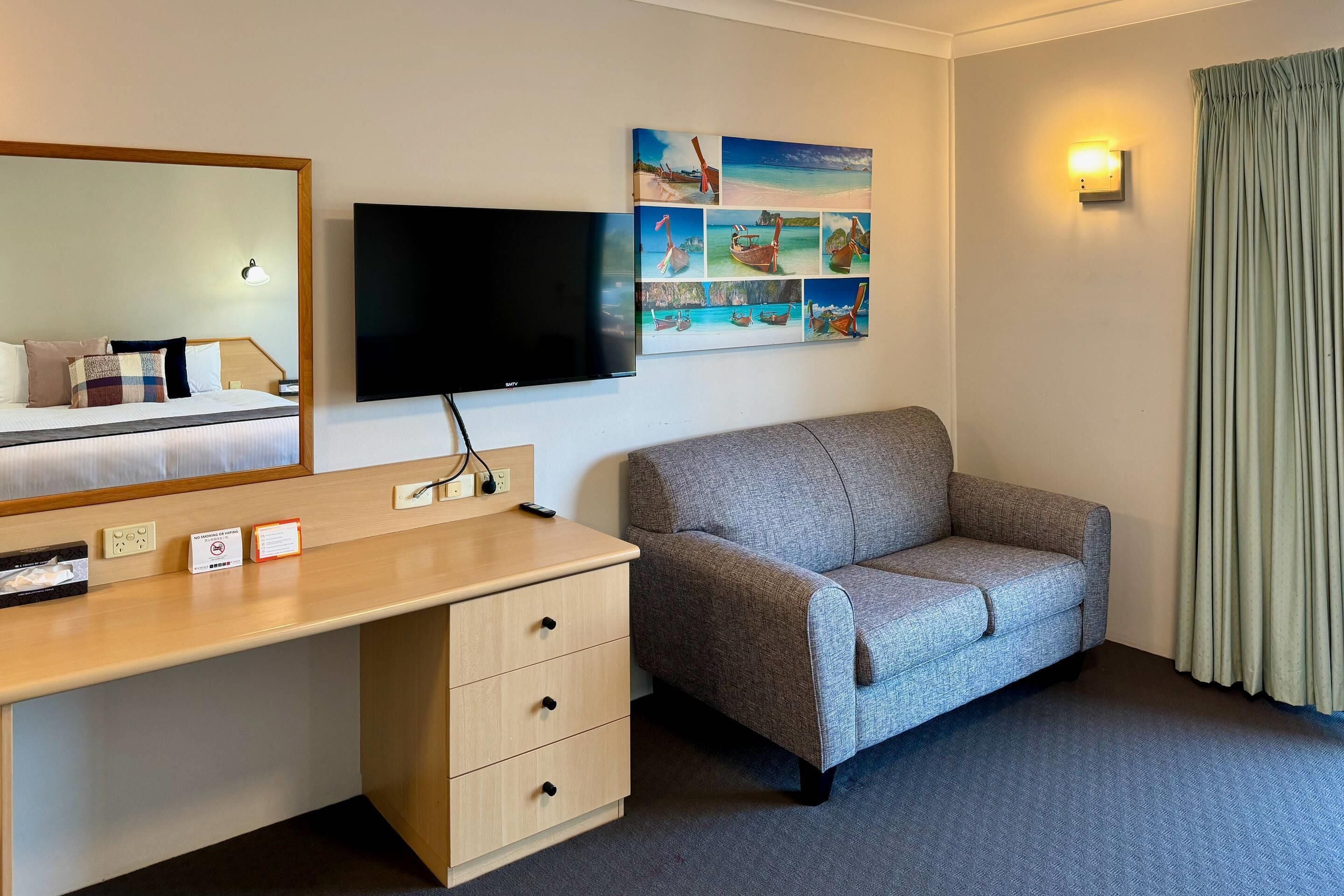 A hotel room with a bed, a desk with drawers, a flat-screen TV, a grey sofa, a wall-mounted picture, and a window with curtains.